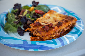 A meal of lasagna and a side green salad on a disposable paper plate
