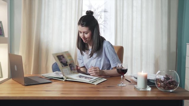 A woman sitting at a table at home The girl is thinking about family and look through photo album. Woman drinks wine and relax. Remote work. Home office. Self-isolation. Quarantine. Epidemic.