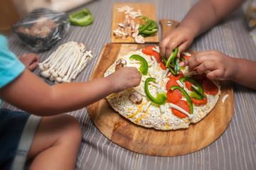 Children adding fresh toppings to a frozen cheese pizza on a wooden pizza peel.
