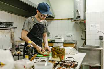 Man with glasses and a cap cooking in a restaurant kitchen with pots of food, oil and pans
