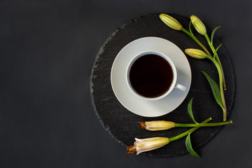 Cup of black coffee tea and fresh spring buds of lily flowers on a round slate on a black background. Romantic stylish breakfast. Date on restaurant. Still life flat lay. Top view. Space for text.