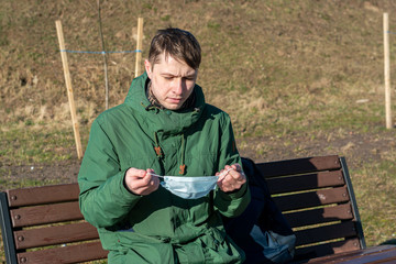 guy is sitting on a bench in the street. puts on a face a white protective medical mask. protection...