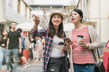 copy space real moments concept. two funny women travelers walking outdoors in summer time holidays bazaar. young girl point aside and showing while friend holding mobile phone searching online map.
