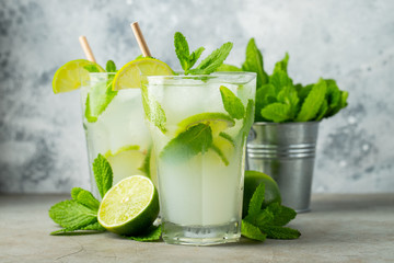 Two homemade lemonade or mojito cocktail with lime, mint and ice cubes in a glass on a light stone...