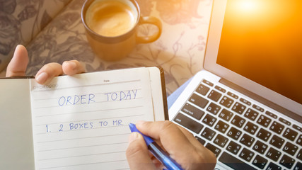 Work space with a laptop. There are a cup of coffee and notebook. A women is holding a notebook and write on "ORDER TODAY 1. 2 Boxes TO Mr.    " , e-commerce concept. 