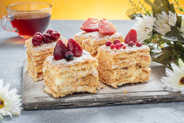Delicious layer cake Napoleon decorated with berries and powdered sugar on a gray stone Board on a gray and yellow background, horizontally