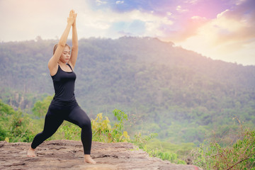 Young woman standing and doing yoga pose at the top of mountain in the morning