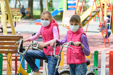 Fototapeta na wymiar Two girls in medical masks ride bicycles in the playground