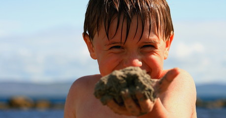 happy boy playing at the beach offeriing sand in his had and smiling
