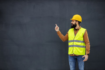 Young smiling bearded worker in vest, with helmet on head standing in front of gray wall and pointing up.