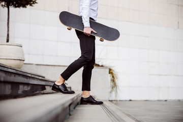 Cropped picture of handsome man in shirt going down the stairs outdoors and holding skateboard...