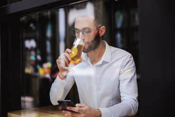 Young businessman dressed in shirt sitting in pub, drinking beer and using smart phone for replying messages on smart phone.