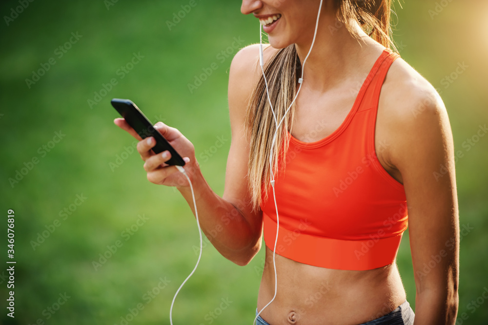 Wall mural Cropped picture of young slim charming sportswoman standing in nature, having earphones in ears and using smart phone for choosing music.