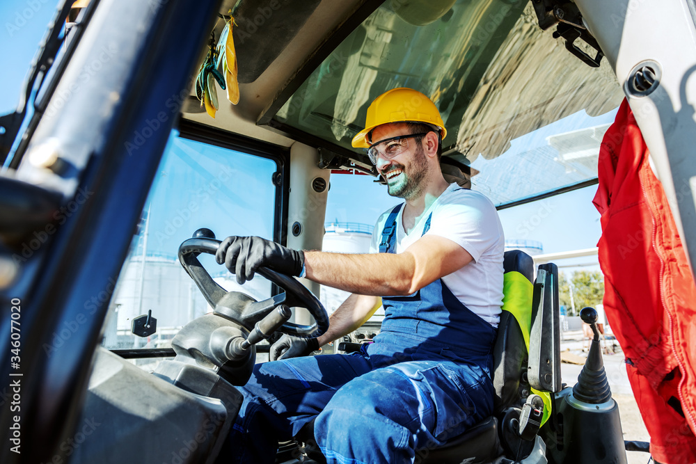 Wall mural smiling handsome caucasian worker in overall and with helmet on head driving excavator.