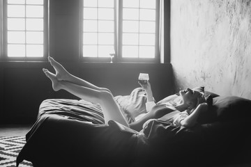 Lonely beautiful woman in the bedroom with large windows on the bed with a glass of wine relaxes. Black and white art photo in a low key. Soft selective focus.