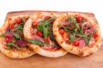 Three pizzas with ham, tomatoes, arugula on a wooden tray.