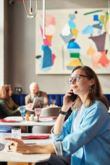 Content confident lady entrepreneur in eyeglasses sitting at table in art cafe and talking on mobile phone