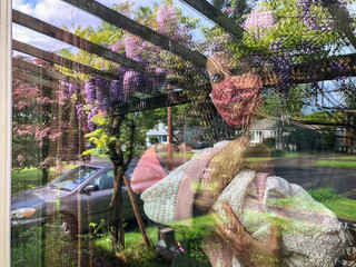 A woman wearing a mask and holding a newborn baby look out of a window in a residential area.  Blooming garden flowers are reflected in the window.