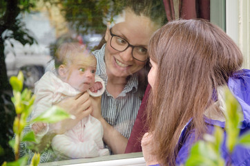 A young mother introduces her newborn baby to family from behind the protection of a window to prevent the spread of the coronavirus.  Three generations of woman are visible.