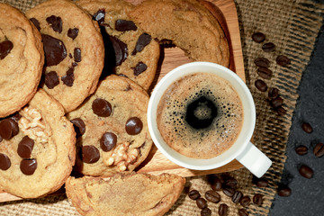 Top view of hot fresh coffee in a white cup with foam and with homemade less sugar chocolate cookies and coffee beans on black table background. Drink and Desserts concept. Dark tone.