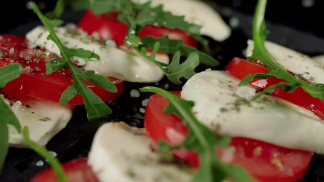 Camera moves up above ready-made caprese salad. Fresh juicy tomatoes, mozzarella cheese and arugula leaves on a black board with a Mediterranean herbs and sea salt. Side view. Close up