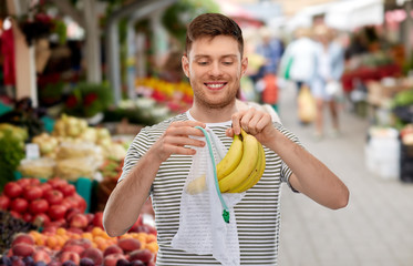 food, sustainability and eco living concept - smiling young man in striped t-shirt putting bananas into reusable string bag over street market on background