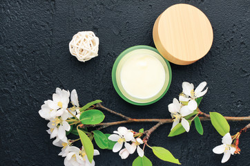 Fototapeta na wymiar Botanical spa cosmetic body care product in green jar with fresh flower blossom, top view black background. Herbal skincare beauty treatment.