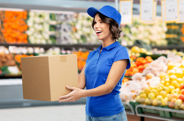 food, service and shipment concept - happy smiling delivery girl with box in blue uniform over supermarket or grocery store on background
