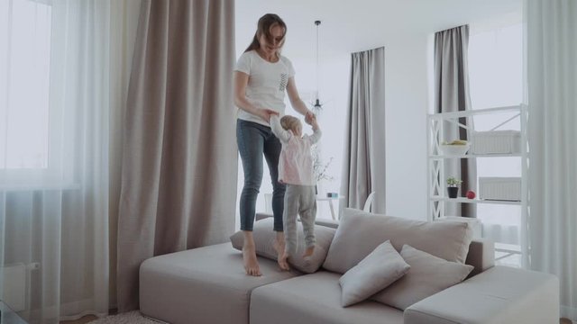 Happy mommy and kid daughter holding hands jumping on sofa together, baby sitter or mother playing having fun with cute kid girl at home, young mum and child enjoy spending time laughing together
