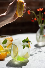 Woman's hand squeezing lemon juice into a glass with lemonade with oranges, lemons and peppermint. Sunny day with fresh beverage on a table