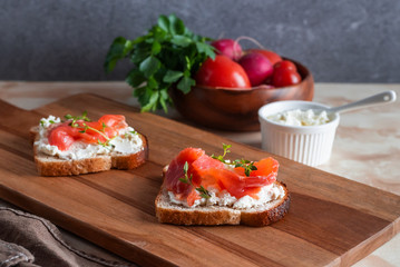 Sandwiches from bread toasts with cereals and bran, cottage cheese and red fish. Close-up of two slices of delicious snacks lie on a wooden cutting board, bowl of vegetables on a marble table.