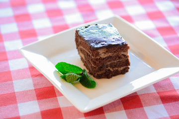 Chocolate brownie piece on white plate decorated with mint leaves served on a white plate over red plaid tablecloth.