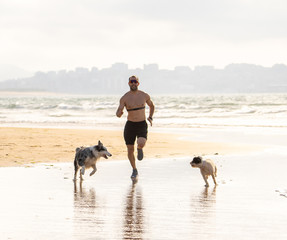 Sport man running with dogs and workout with heart rate monitor on dog-friendly beach. Back to Life Outdoors training after coronavirus restrictions and lockdown easing.