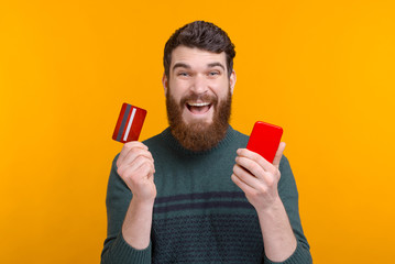 Excited bearded man is showing his card and phone on yellow background. Happy to use mobile banking.