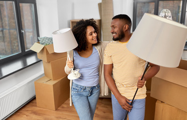 Fototapeta na wymiar moving, people, repair and real estate concept - happy african american couple with lamps packing stuff into cardboard boxes at home