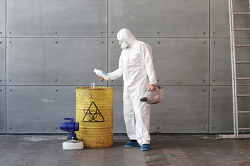 a man in a protective suit and mask examines bottles next to a yellow barrel