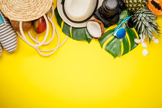 Straw hat, camera, bag, summer shoes, sunglasses, shells, tropical leaves and fruits over yellow background