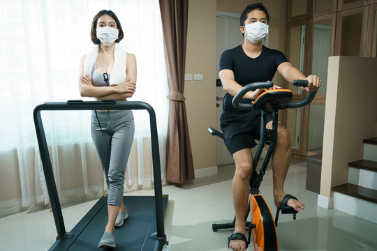 Exercise At Home: Man Biking Exercise Or Indoor Cycling And Woman Running On A Treadmill With Mask : Health Care During Coronavirus Pandemic.