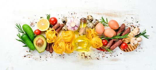 Food background. Dry pasta, tomatoes, spices and vegetables on a white wooden background. Top view.