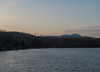 Evening view on calm water of forest lake, fish pond Kunraticky rybnik with birch and spruce trees growing along the shore and Fisherman angler. Pink sunset sky with clouds.