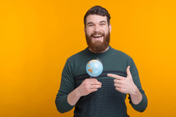 Young bearded male student is pointing at his small globe over yellow background.