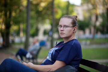 A woman in blue jeans and a blue T-shirt sits on a bench in the park and listens to music. Red headphones. Girl in glasses.