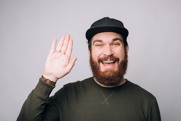 Excited bearded man smiling at the camera is making hello gesture with one hand on white background. © Vulp