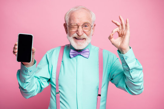 Portrait of energetic positive old man hold new cellphone show okay sign recommend choose good modern technology wear teal turquoise shirt violet bow tie isolated pastel pink color background