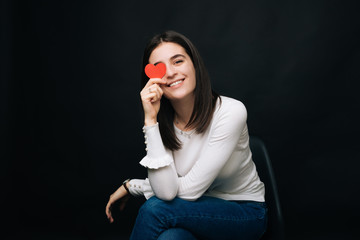 Cheerful young woman sitting on a chair is covering one eye with a red heart on black background.