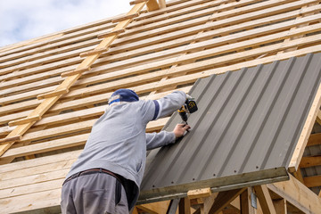 Senior gray-haired Construction man using a screwdriver, fastens a roofing sheet to wooden rafters on the roof of a country house under construction. Physical activity of the seniors. - obrazy, fototapety, plakaty