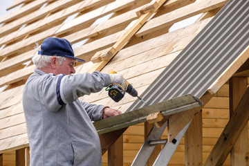 Senior gray-haired Construction man using a screwdriver, fastens a roofing sheet to wooden rafters...