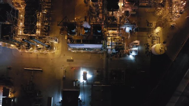 Night aerial view over petroleum refinery with smoking chimneys and many steel technical constructions brightly lit with lights. Smoke rises up into black sky. Environment polluting manufacturing. 4K