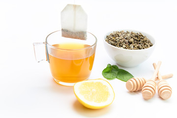 Hot cup of tea with lemon and mint leaves next to a bowl of green tea leaves on a white background