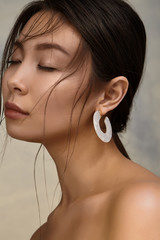 Cropped portrait shot of a young Asian woman with naked shoulders and closed eyes. The lady is wearing a stud earring in the shape of a disc segment and made of white mica-like material with sparkles.